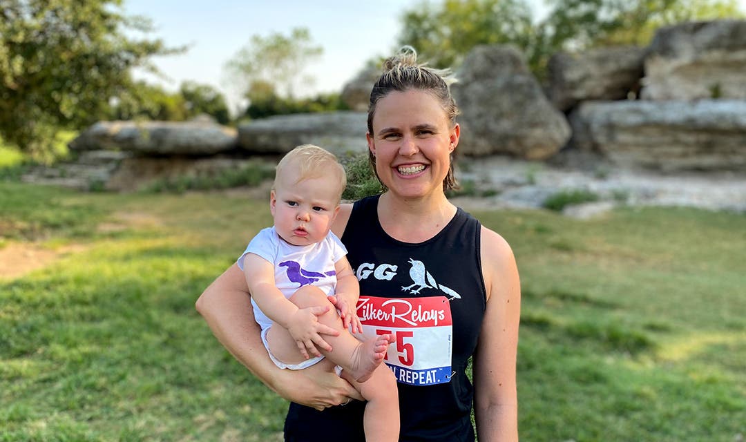 Cate Barrett smiling for the camera wearing a race bib and holding her infant. 