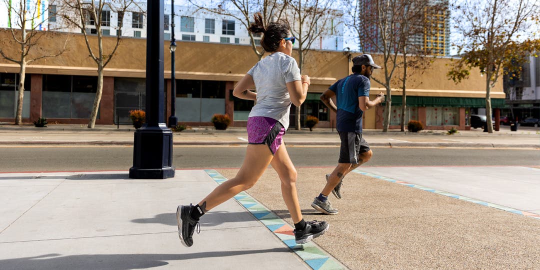 A man and woman running together along a wide sidewalk in a semi-urban neighborhood on a sunny day.