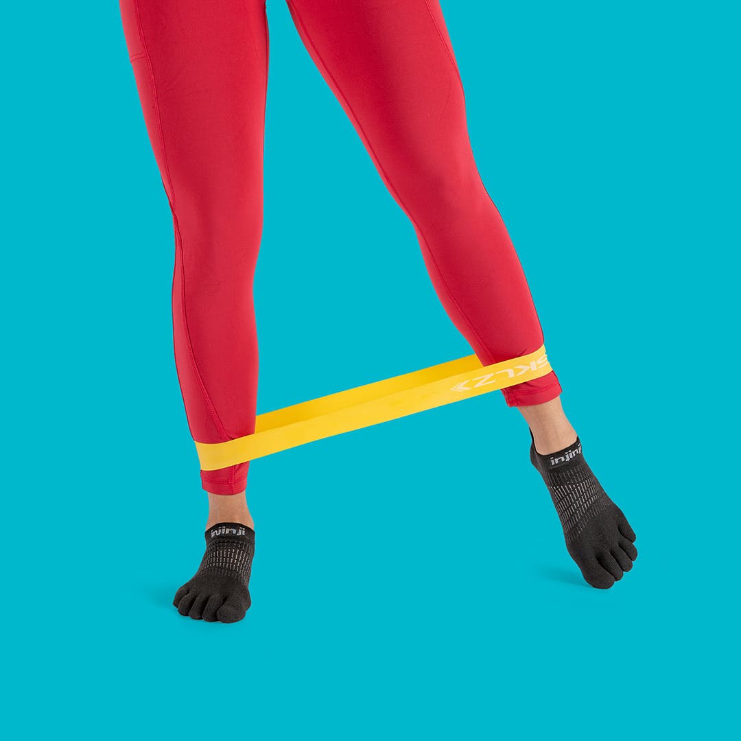 A woman stading upright wearing red leggings and black Injinji Women's Run Lightweight No-Show toesocks while using a resistance band around her ankles. 