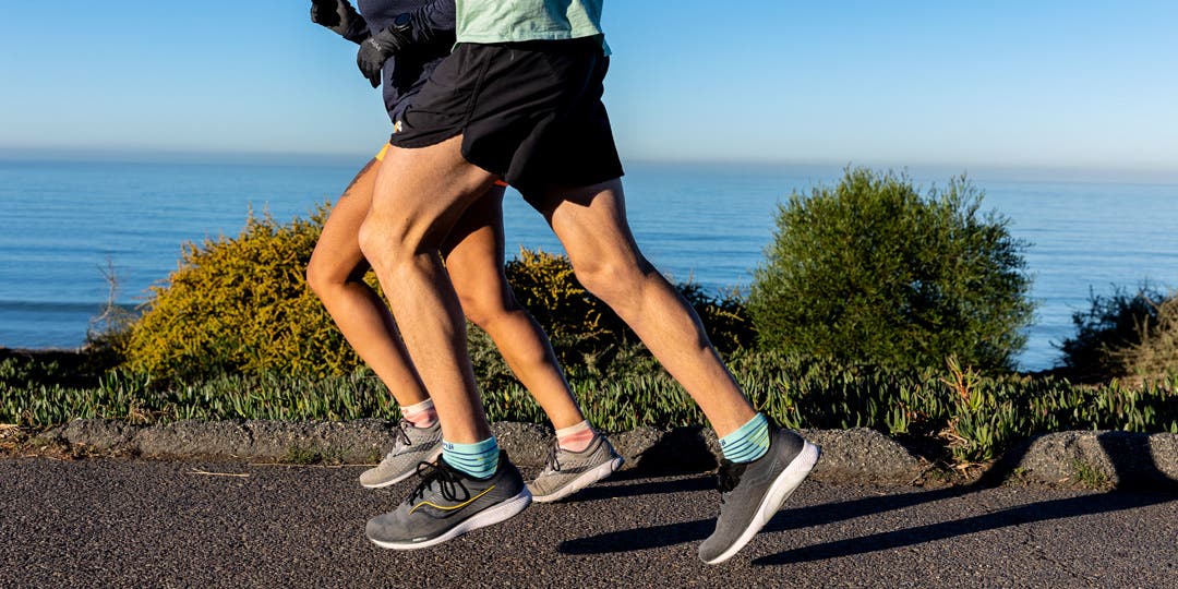 A man and woman running on a paved path on the coast of San Diego with the ocean and blue skies in the background. They're wearing shorts and tees, running shoes, and Injinji Ultra Run Mini-Crew toesocks. 