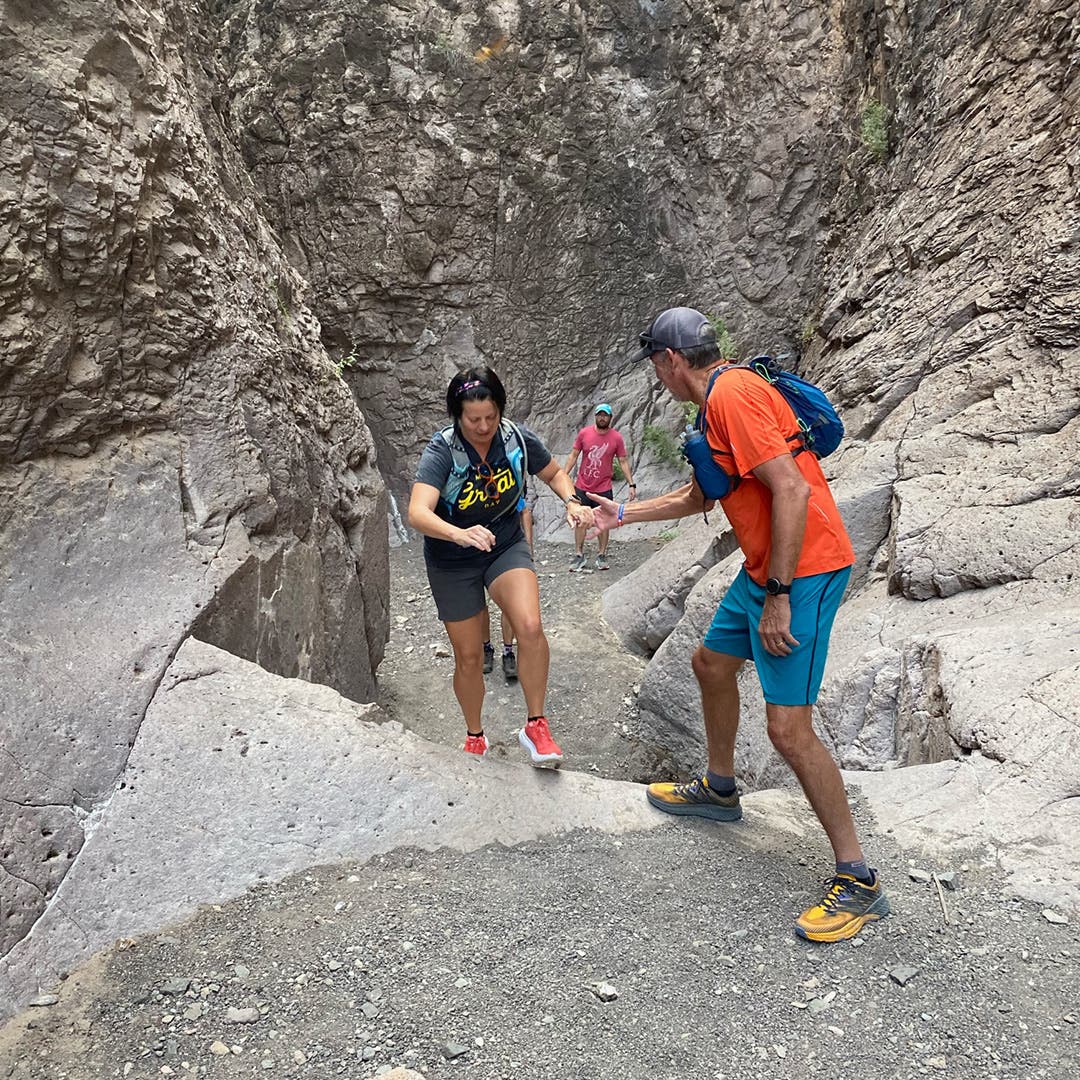 Completely surrounded by large granite walls and boulders, a man helps a woman up a granite stair, with other hikers in the background. 