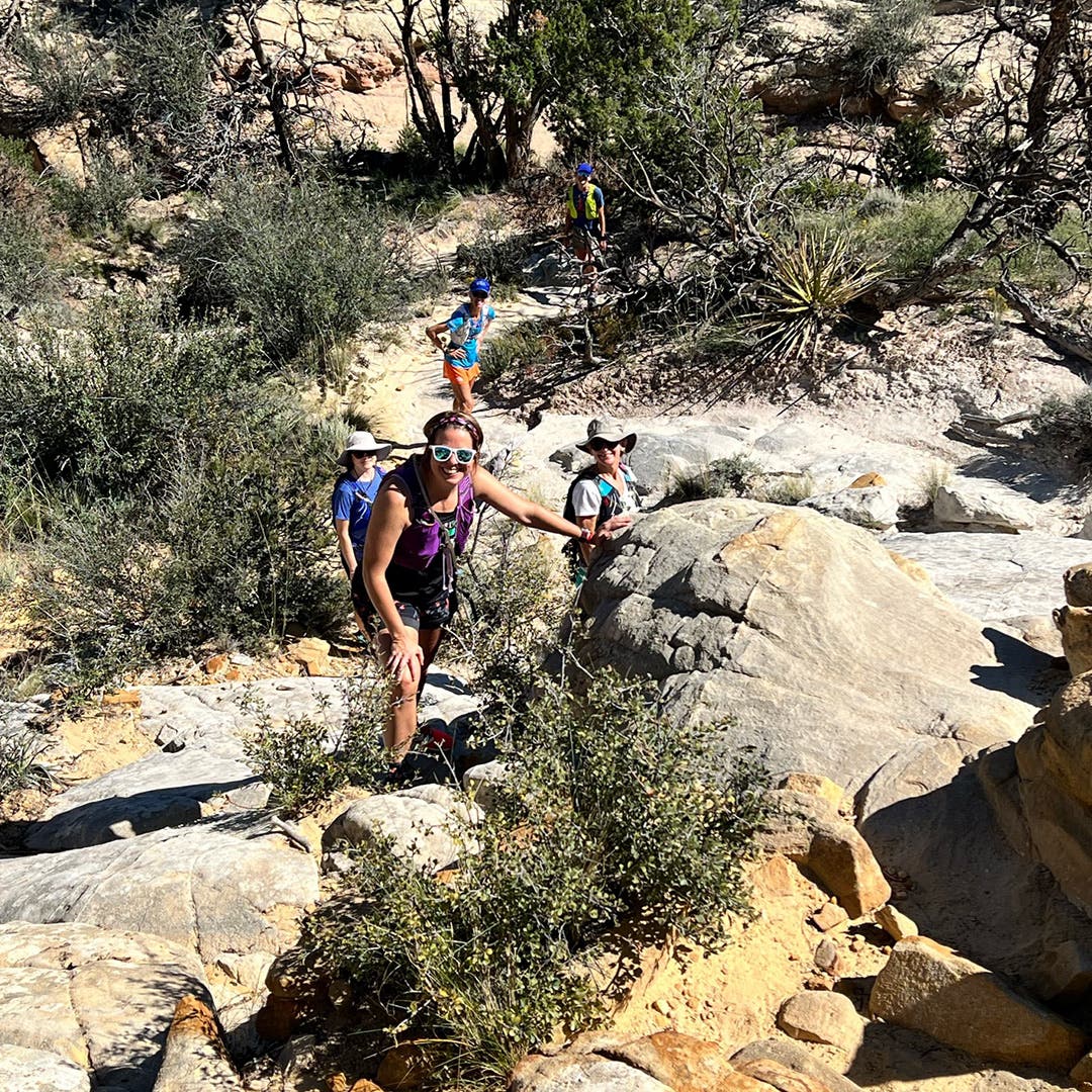 Looking down on a group of hikers from above. They're smiling and turned toward the camera, wearing sunglasses and sunhats in the dusty, high desert landscape.