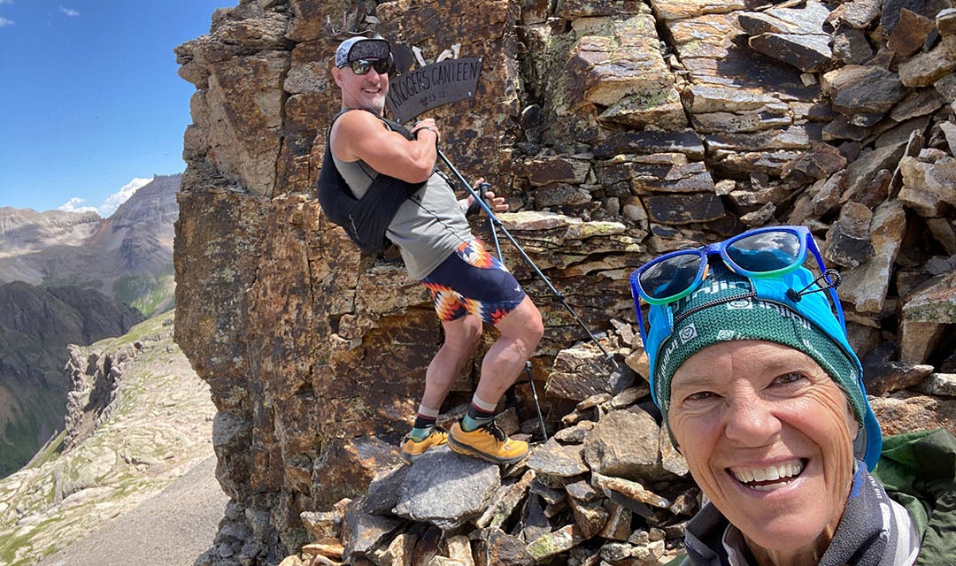 Tanned and happy Ultra runner, Meghan Canfield, smiles at the camera. Behind her is a male hiker standing on a pile of granite stones, smiling and pointing to a sign that says "Krogers Canteen".