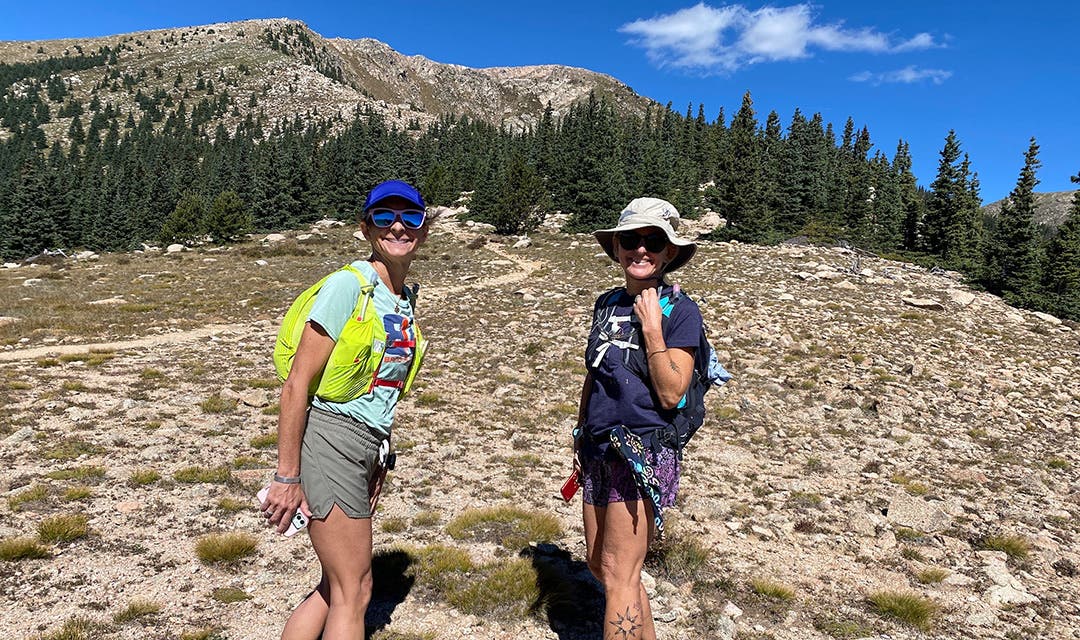 Two female hikers turned and smiling toward the camera among the high desert, alpine landscape of shrubbery and pine trees. 