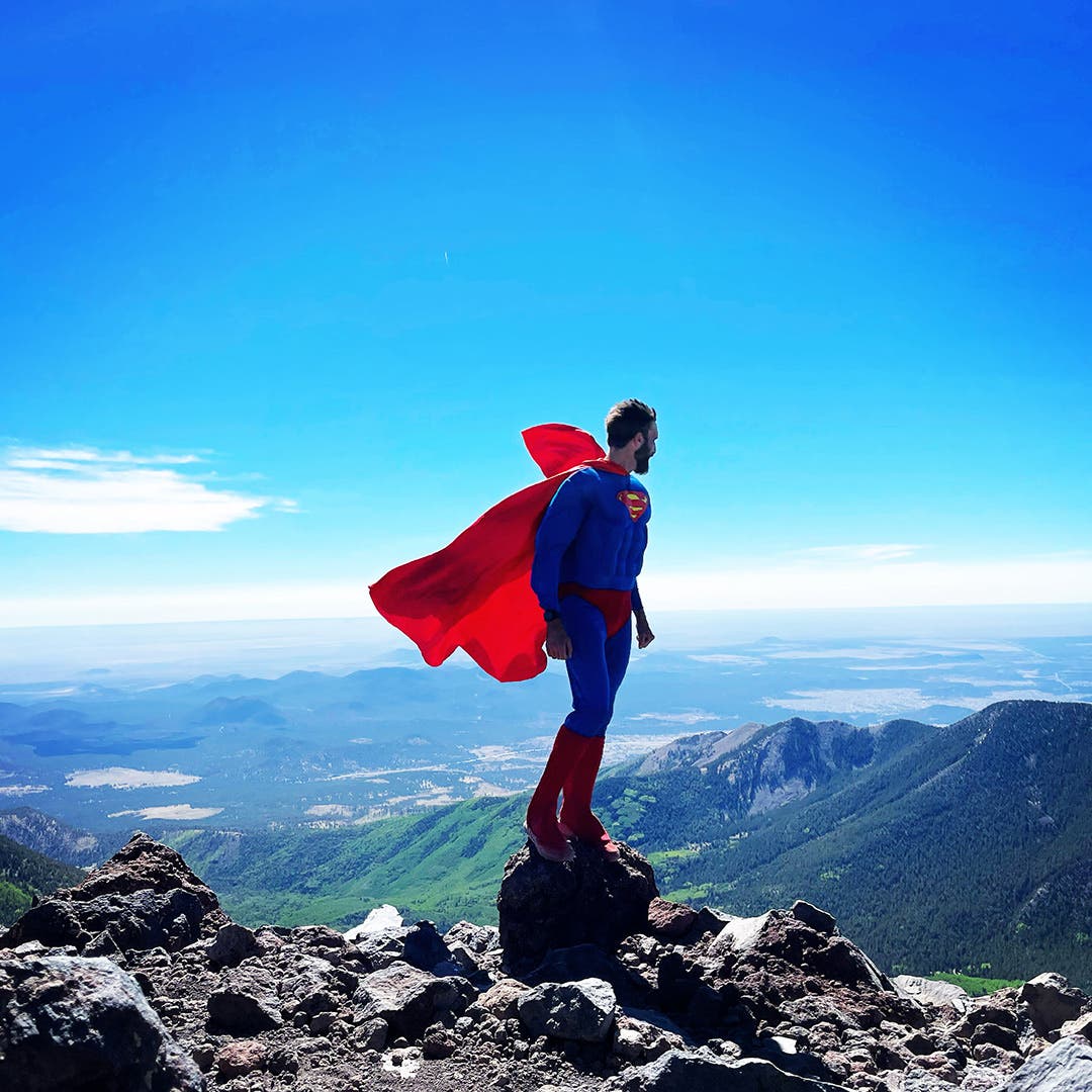 Peter Mortimer standing on a rock on a mountain wearing a superman costume and looking down into the valley below.