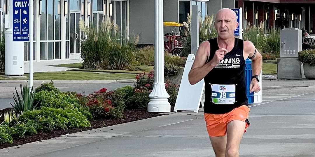 Male ultra athlete in a sprint down a road next to a long white building.