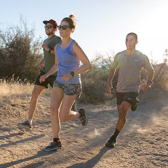 Three pepole running on a sandy, mostly flat trail among desert shrubs.