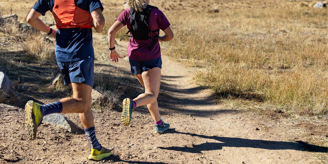 A man and woman running on a dirt trail in the dry mountains of San Diego. They're sporting Trail Midweight Crew and Mini-Crew Injinji socks, hydration vests, shorts and tees. 