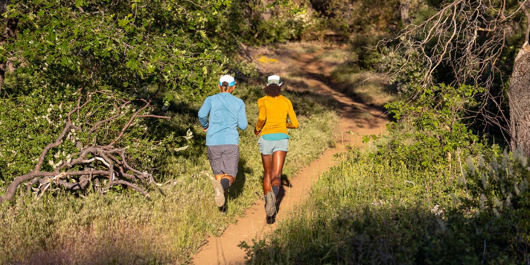 A man and woman running side by side on semi-shaded dirt trail among green trees and shrubs. They're wearing hats, long sleeve tees, shorts, running shoes, and Injinji Trail Midweight Mini-Crew toesocks.
