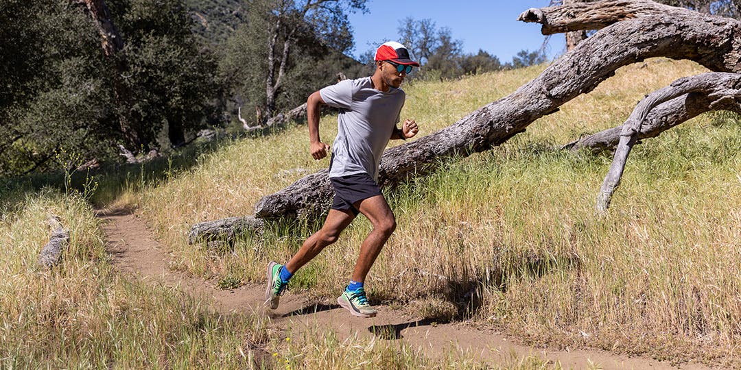 A man running on a dirt trail surrounded by trees and long dry grass.