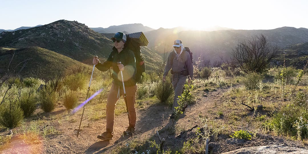 Two backpackers hiking up a dirt trail with mountains in the distance.