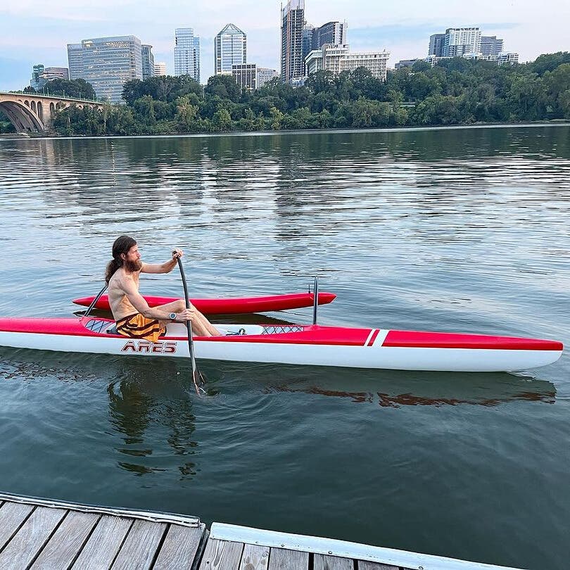 Mike Wardian sitting in a long rowing boat in the river with a big city skyline in the background.