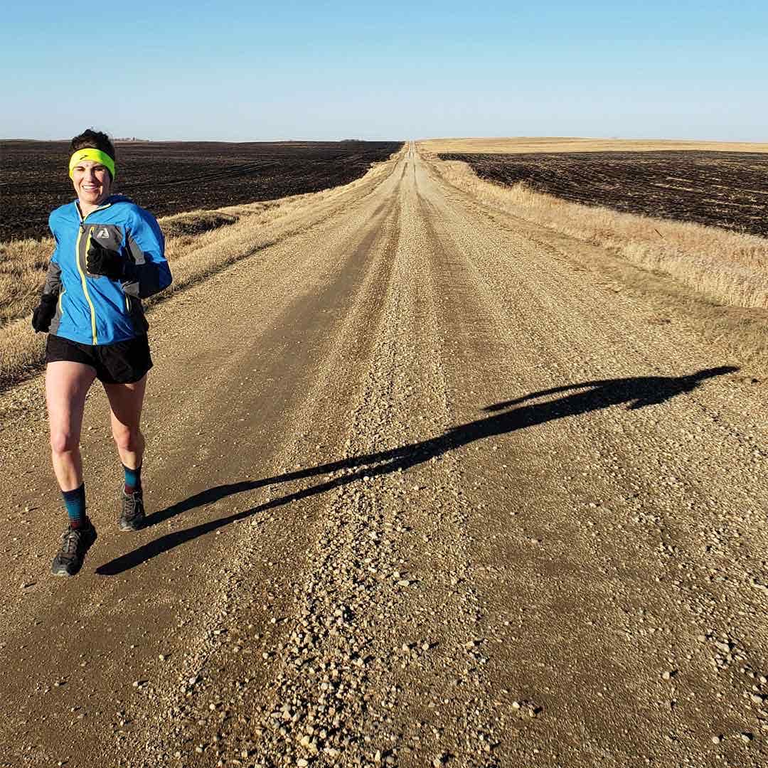 Female running down a dirt road wearing a blue windbreaker, black shorts, neon green earband and gloves. 