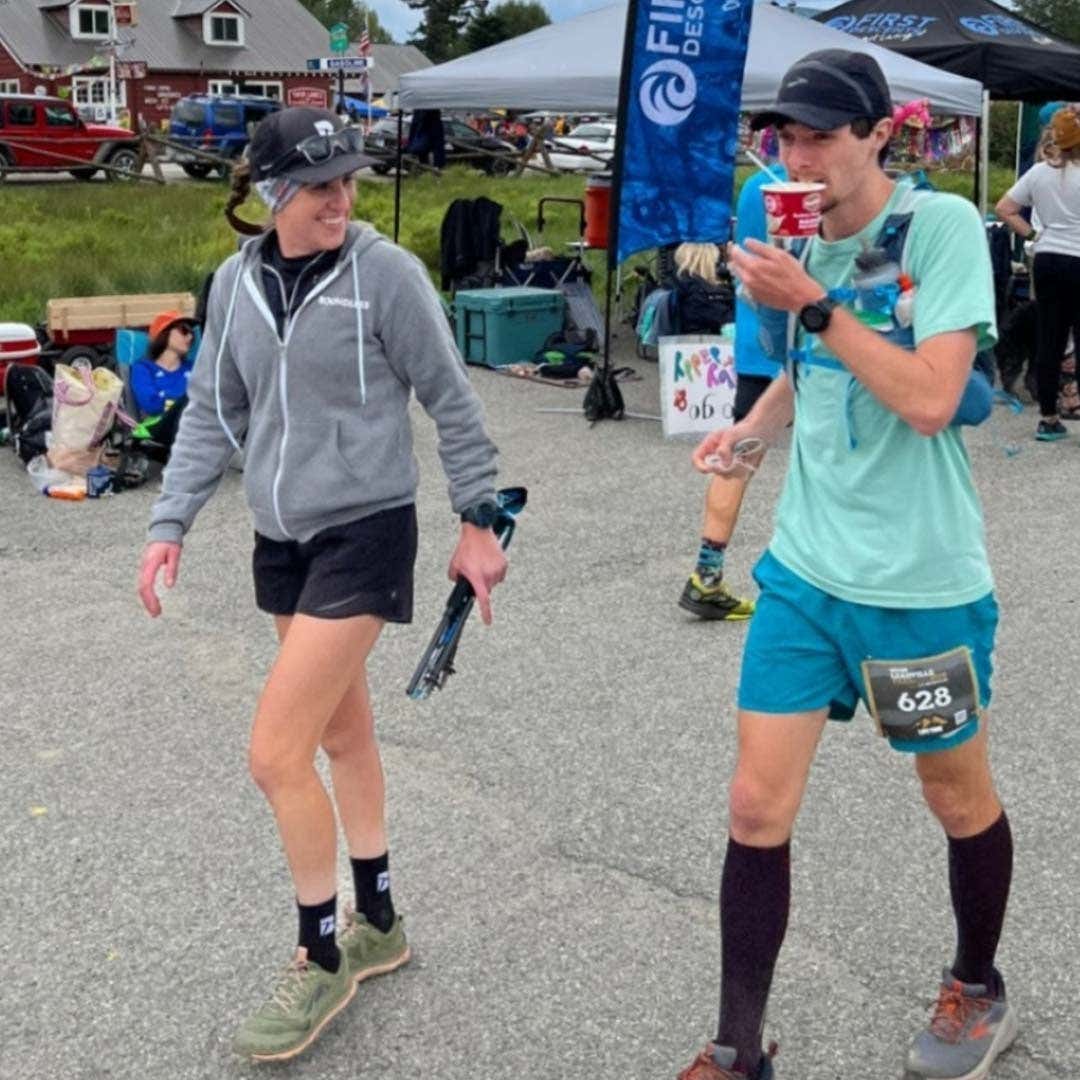 A female wearing a grey hoodie and black shorts with a male wearing a blue shirt and shorts, walking side by side at a race. 