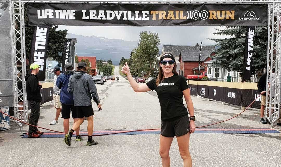 Female athlete wearing a black tee shirt and black shorts with trail shoes, standing in front of a race banner, giving a thumbs up.