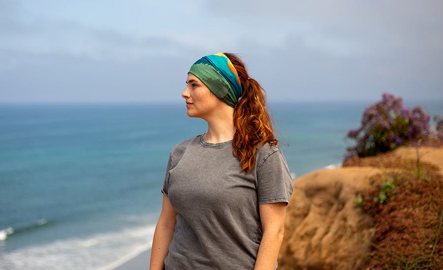 A woman standing on a cliff by the ocean wearing Injinji's Multifunctional Headband Buff in Rocky.