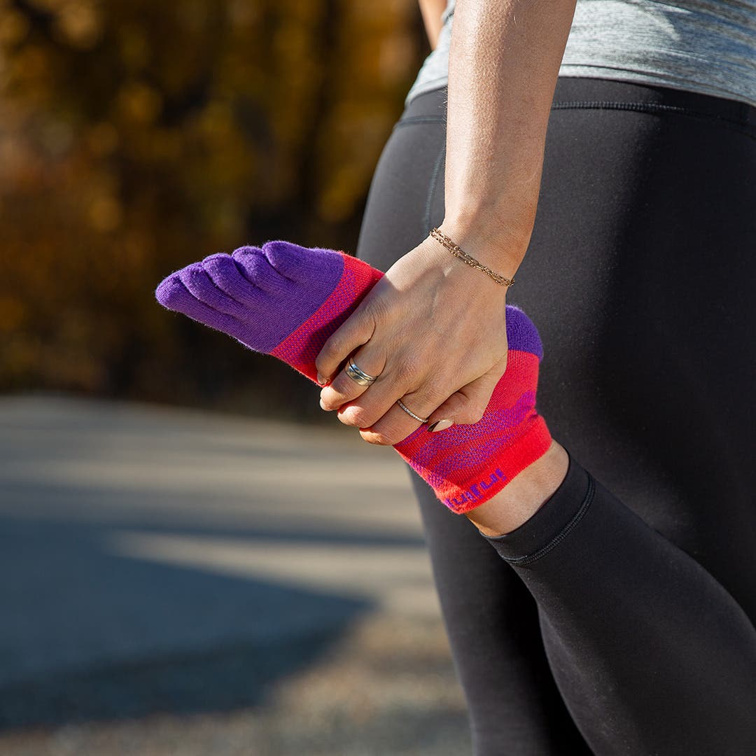 A woman stretching her quads by holding her foot behind her, showing off her Injinji Women's Ultra Run No-Show socks in Razzmatazz.