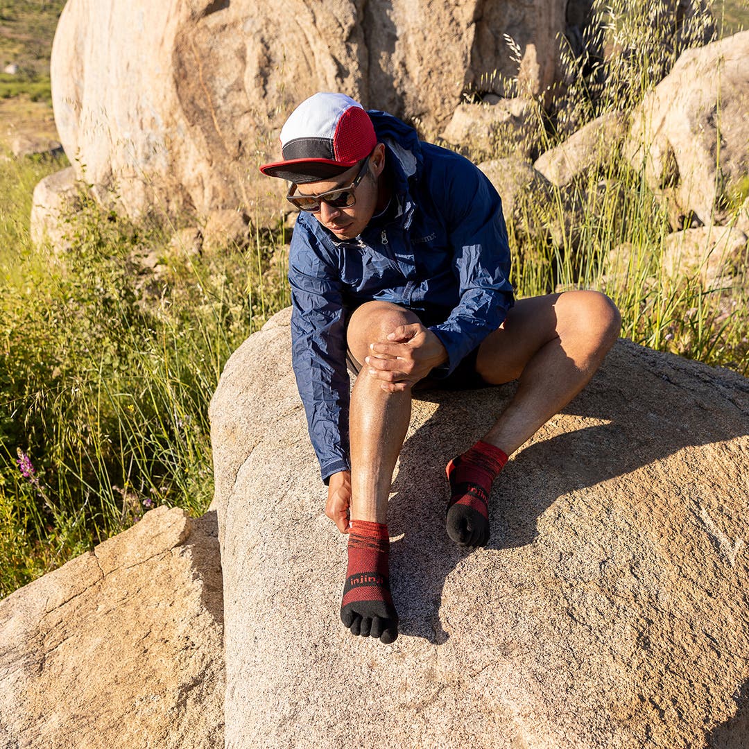 A person sitting on a boulder next to a trail adjusting their Injinji Trail Midweight Mini-Crew socks in Meteor. 