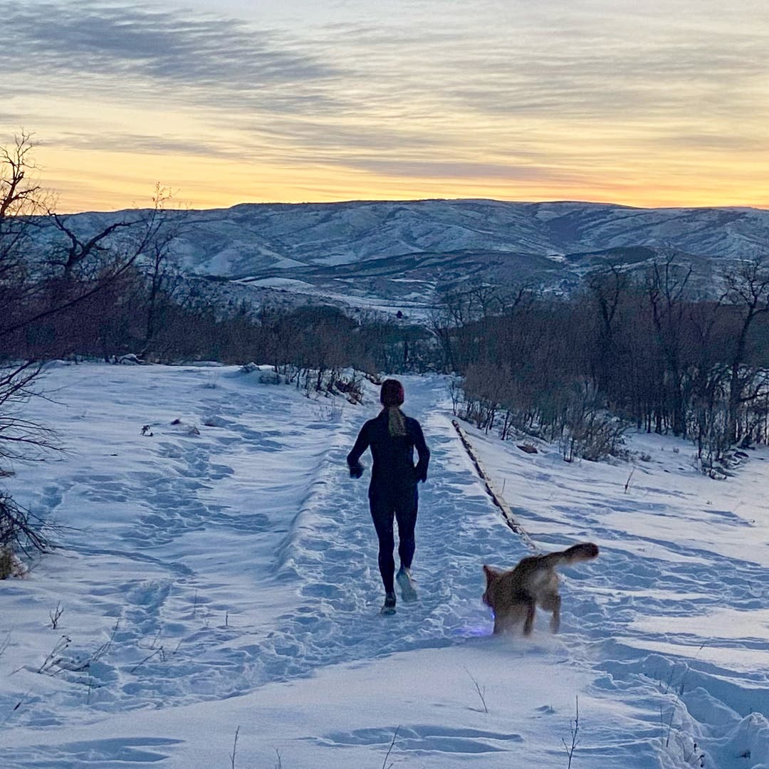 Woman running down a snowy path with her dog at dusk.